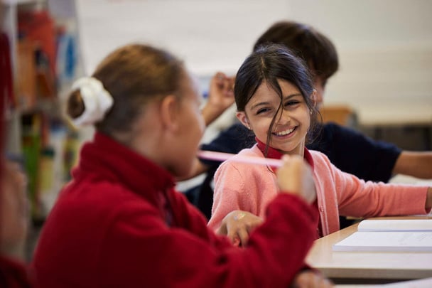 Two girl elementary students chatting and having fun in a class with a boy student in the background.