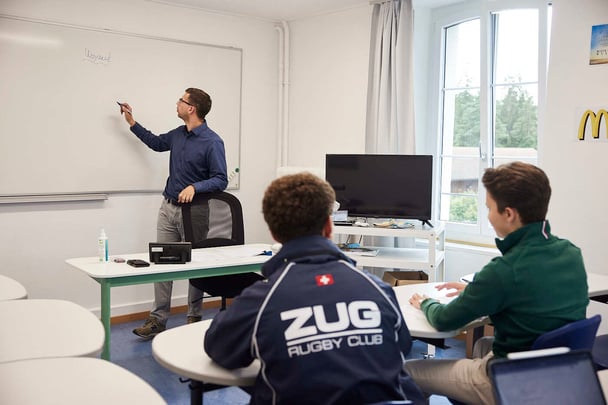 A male teacher is writing something on a white board and two boy students pay attention.