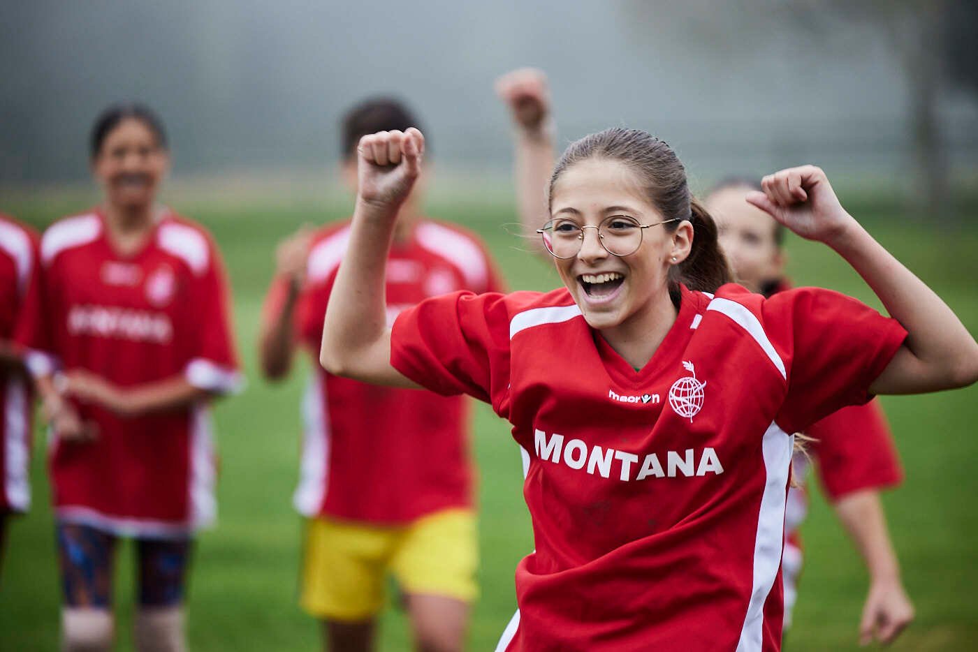 A girl student cheers victoriously on a football field with her cheering friends in the background.