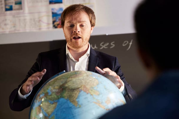 A teacher speaks behind a globe in a classroom.