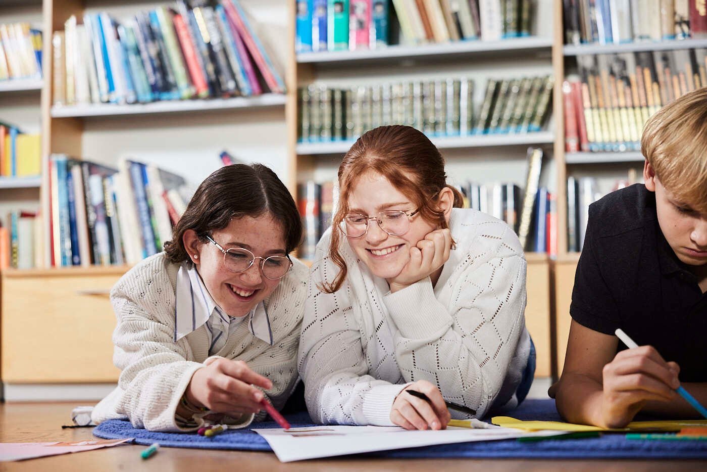 Two girls lying on a floor in a library looking at a paper happily.