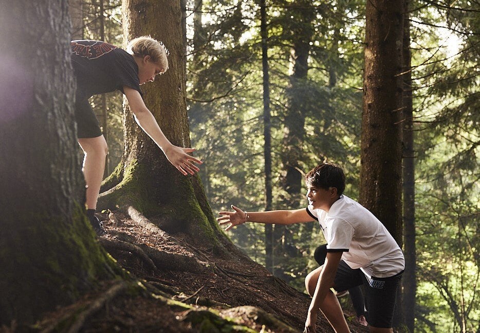 A boy student is extends his arm to help another boy student in a forest.