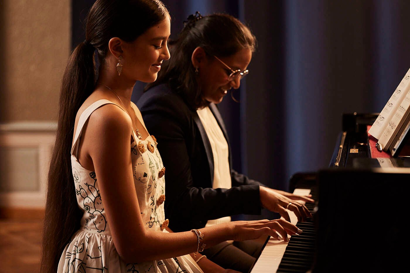 A girl is playing a piano supported by an elder woman next to her.