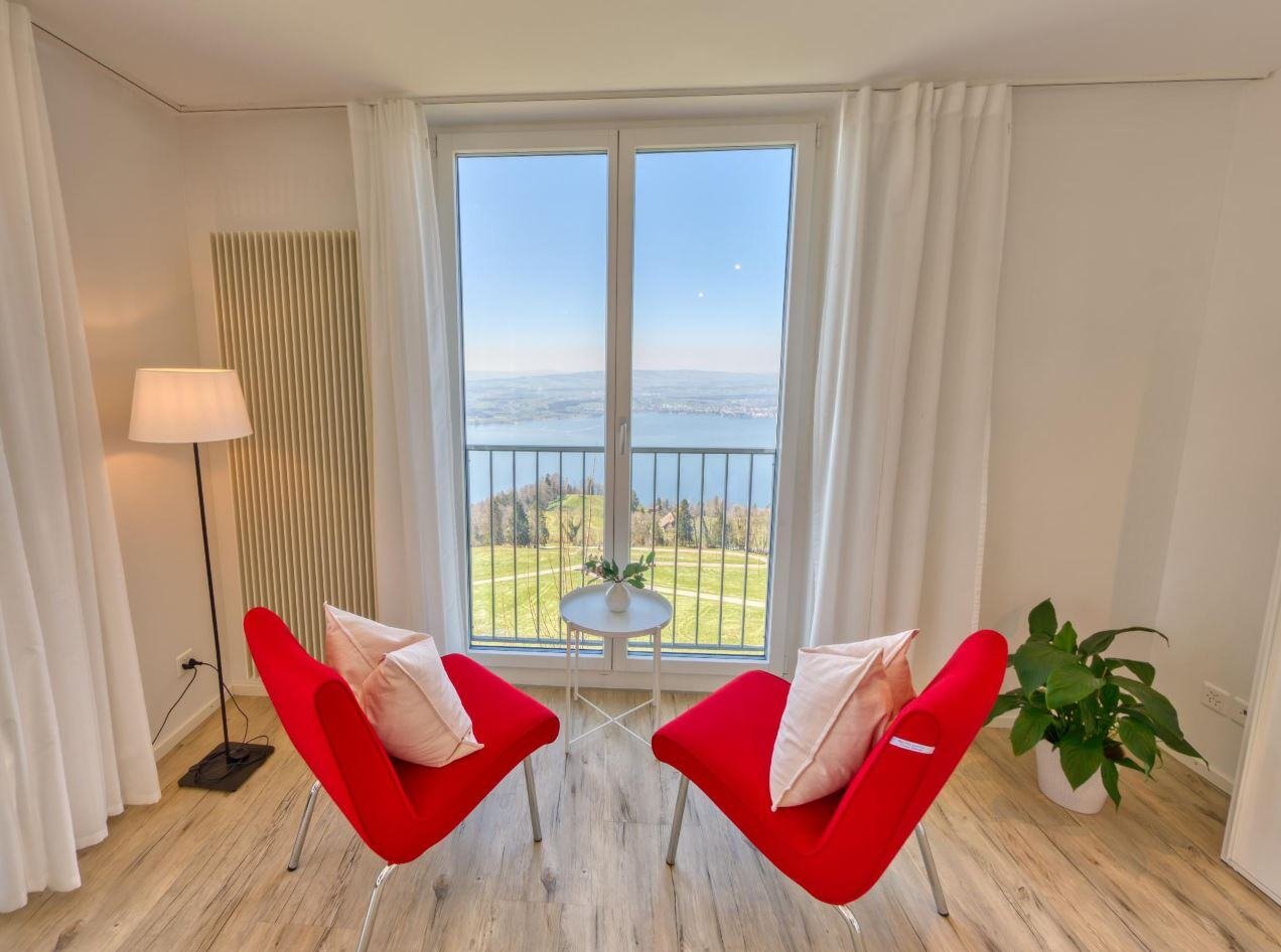 An open window of a bright airy room overlooking the Zug lake view, two red sofas with a small table in front of the window.