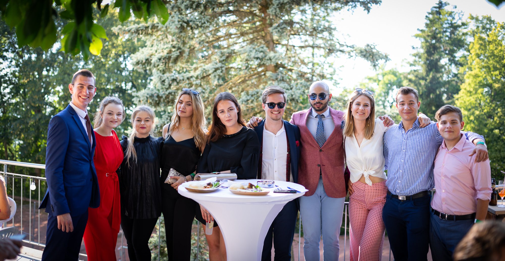 A group of students in formal outfits pose happily to camera in an outdoor setting.