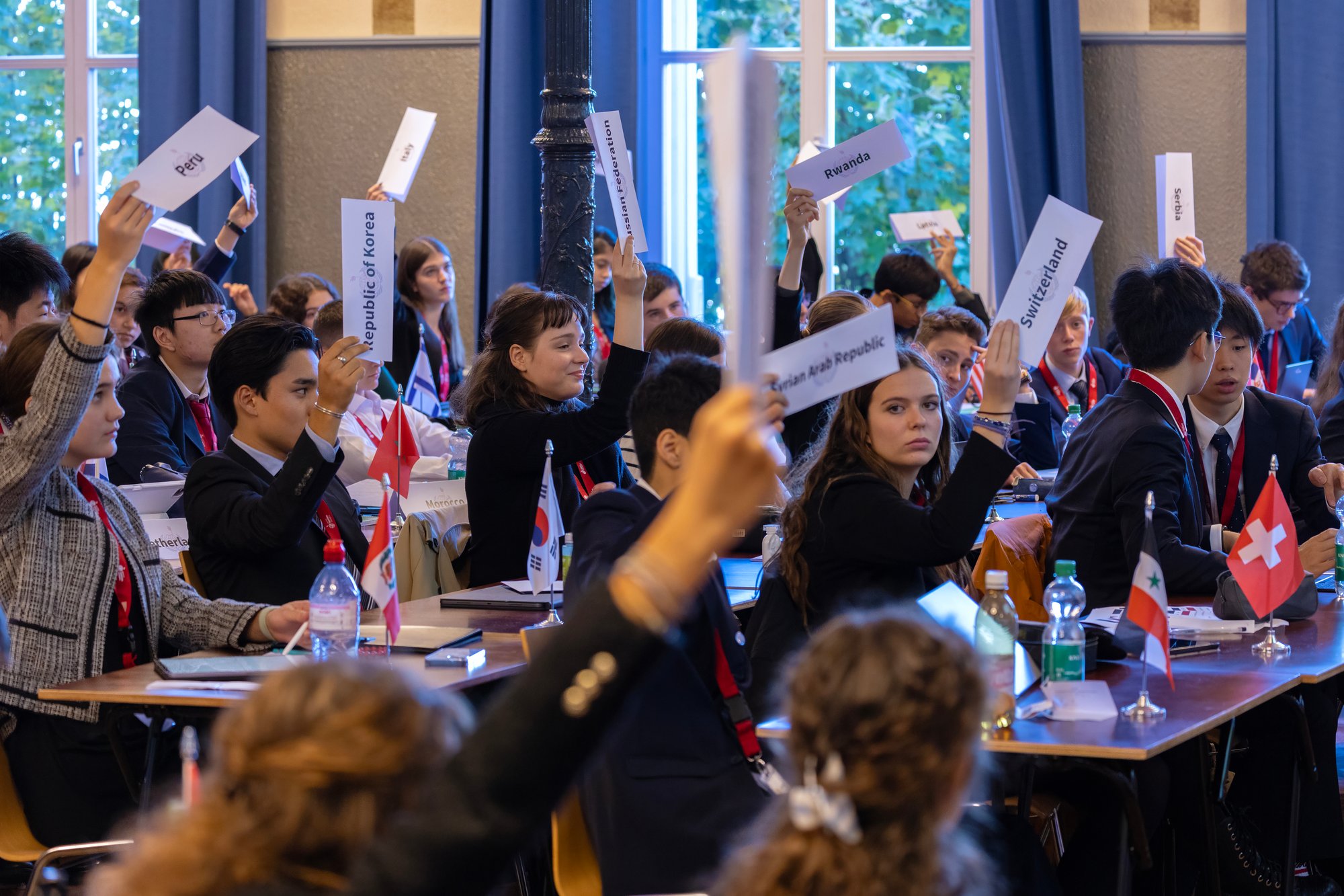 A group of students in the middle of a conference, most of them hold a paper showing their country origin.