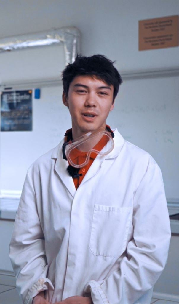 A young mentor stands in a science lab and talks to camera. He wears a science robe and the work glasses.