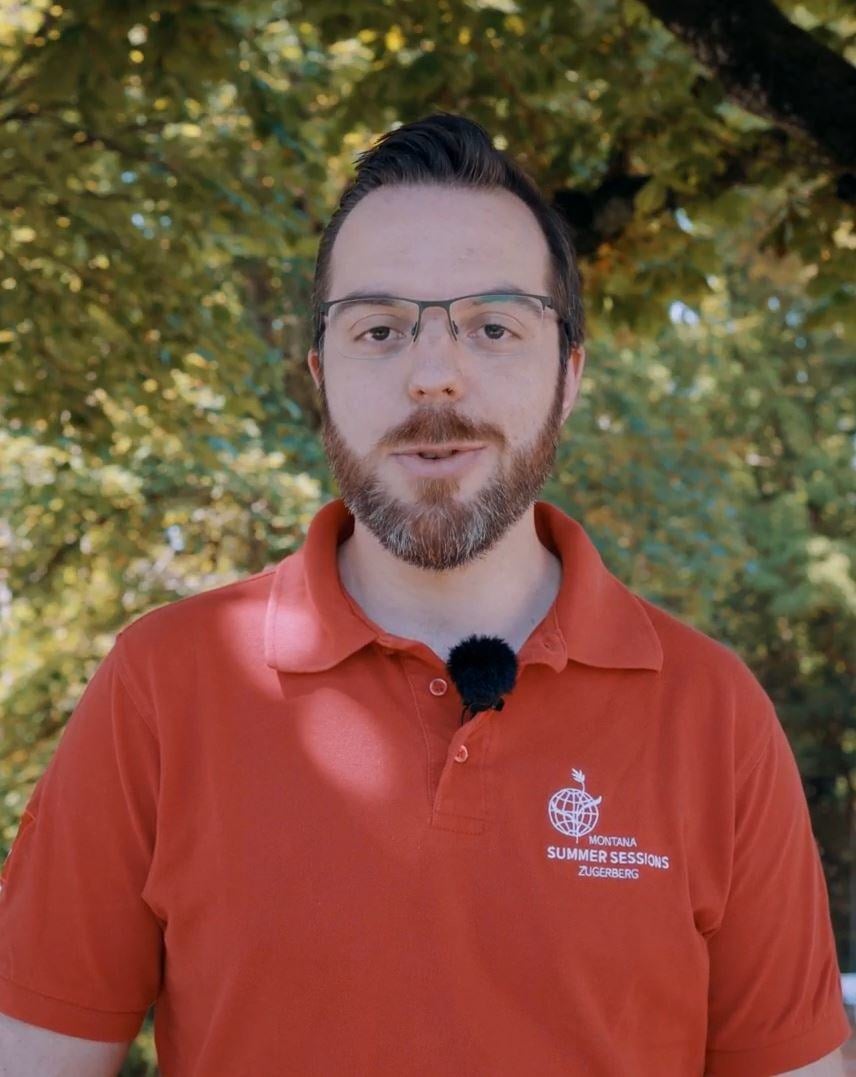 A male staff talks to camera. He wears a red top written Montana Summer Sessions and also glasses.