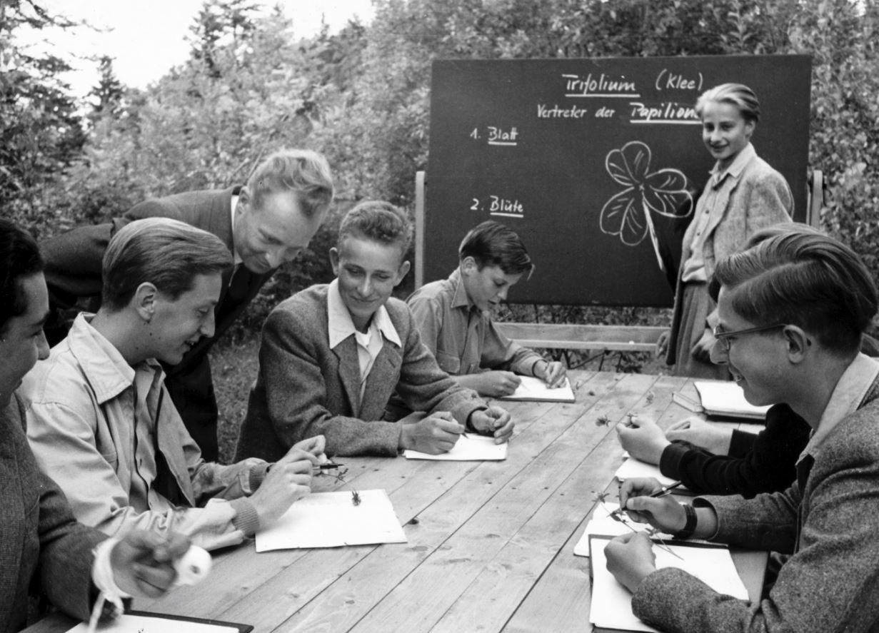 Several male students studying outside with a teacher and an elder woman.