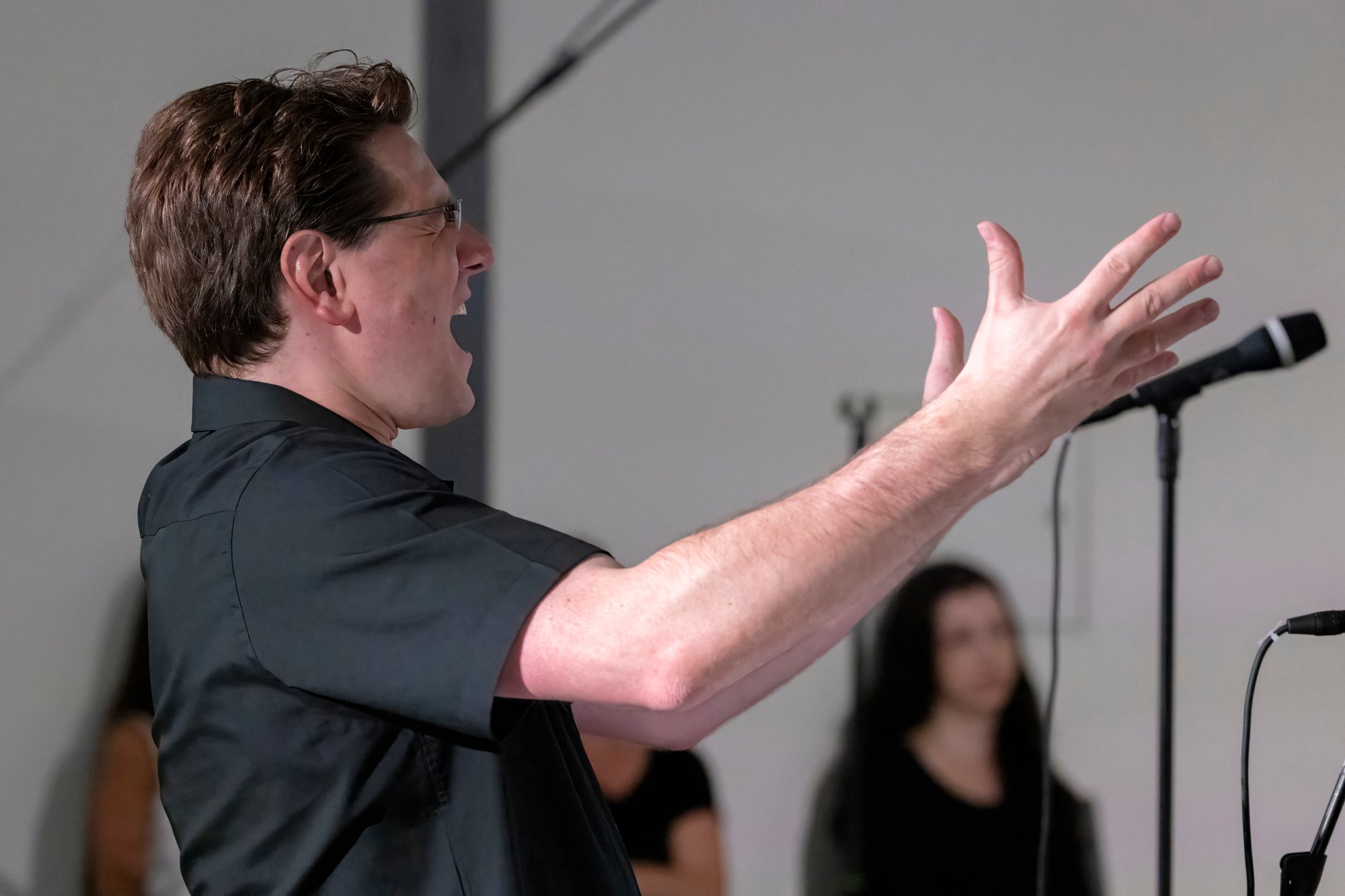 A man in black outfit conducts a choir in a room.