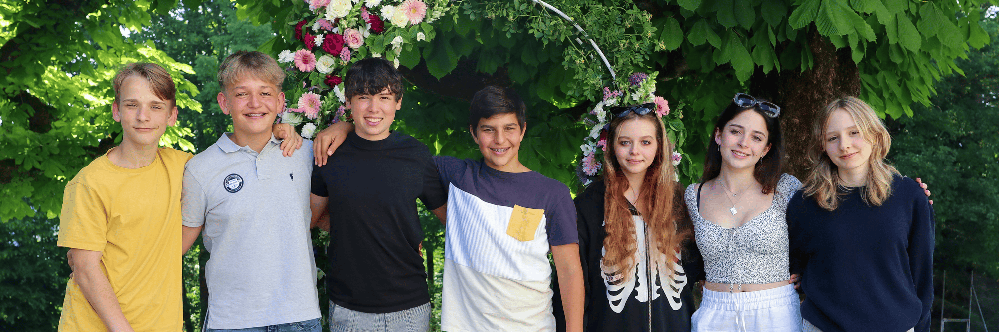 Seven young students of boys and girls pose in a bushy garden.